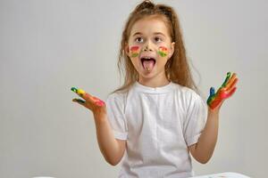 Little girl in white t-shirt is posing standing isolated on white, showing her pained hands and tongue. Art studio. Close-up. photo