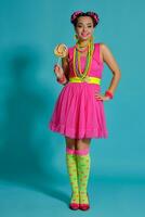 encantador niña con un multicolor trenzas peinado y brillante constituir, posando en estudio en contra un azul fondo, participación un pirulí en su mano. foto