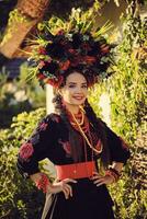 morena niña en negro y rojo bordado ucranio auténtico nacional disfraz y un guirnalda de flores es posando en pie en contra un blanco cabaña. foto