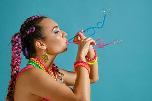 encantador niña con un multicolor trenzas peinado y brillante constituir, es soplo burbujas utilizando túbulos, posando en estudio en contra un azul antecedentes. foto