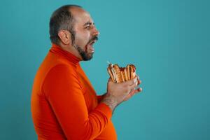 Close-up portrait of a middle-aged man with beard, dressed in a red turtleneck, posing with burgers against a blue background. Fast food. photo