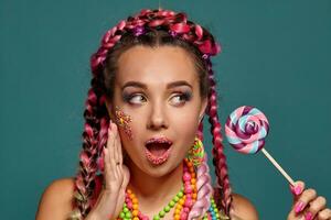 Lovely girl with a multi-colored braids hairstyle and bright make-up, posing in studio against a blue background, holding a lollipop in her hand. photo