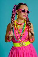 encantador niña con un multicolor trenzas peinado y brillante constituir, posando en estudio en contra un azul fondo, participación un pirulí en su mano. foto