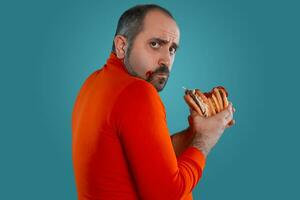 Close-up portrait of a middle-aged man with beard, dressed in a red turtleneck, posing with burgers against a blue background. Fast food. photo