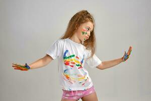 Little girl in white painted t-shirt is posing standing isolated on white and gesticulating with her colored in different paints palms and face. Art studio. Close-up. photo