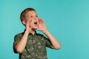 Close-up portrait of a blonde teenage boy in a green shirt with palm print posing against a blue studio background. Concept of sincere emotions. photo