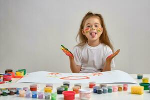 Little girl in white t-shirt sitting at table with whatman and colorful paints on it, showing her pained hands, tongue. Isolated on white. Close-up. photo