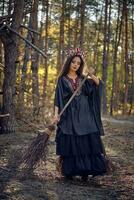 bruja en negro, largo vestido, con rojo corona en su largo cabello. posando con Escoba en pino bosque. hechizos, magia y brujería. lleno longitud retrato. foto
