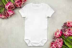 White baby girl or boy bodysuit mockup flat lay with tulip flowers on gray concrete background. Design onesie template, print presentation mock up. Top view. photo