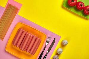 Sausages with tomatoes, spaghetti and garlic on a yellow and pink minimal background. Flat lay. Top view. photo