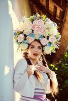 morena niña en un blanco ucranio auténtico nacional disfraz y un guirnalda de flores es posando en contra un blanco cabaña. foto