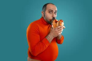 Close-up portrait of a middle-aged man with beard, dressed in a red turtleneck, posing with burgers against a blue background. Fast food. photo