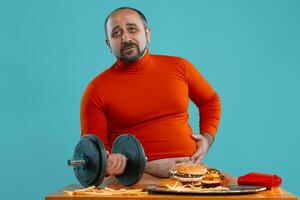 Close-up portrait of a middle-aged man with beard, dressed in a red turtleneck, posing with burgers and french fries. Blue background. Fast food. photo