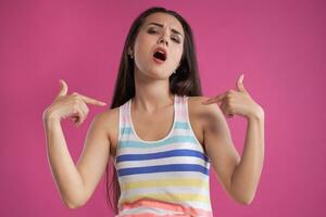 Brunette woman with long hair, dressed in colorful striped shirt, posing against pink studio background. Sincere emotions. Close-up. photo