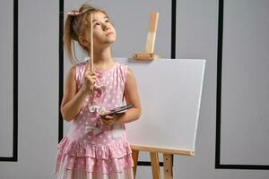 Little girl artist in a pink dress is standing behind easel and painting with brush on canvas at art studio with white walls. Medium close-up shot. photo