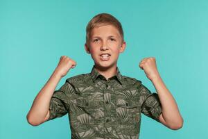 Close-up portrait of a blonde teenage boy in a green shirt with palm print posing against a blue studio background. Concept of sincere emotions. photo