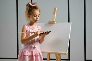 Little girl artist in a pink dress is standing behind easel and painting with brush on canvas at art studio with white walls. Medium close-up shot. photo