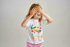 Little girl in white painted t-shirt is posing standing isolated on white and gesticulating with her colored in different paints palms and face. Art studio. Close-up. photo