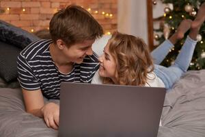 Smiling young couple man and woman lying in bed with laptop and looking at each other photo