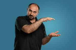 Close-up portrait of a brunet middle-aged man with beard, dressed in a black t-shirt and posing against a blue background. photo