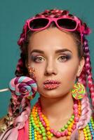 encantador niña con un multicolor trenzas peinado y brillante constituir, posando en estudio en contra un azul fondo, participación un pirulí en su mano. foto