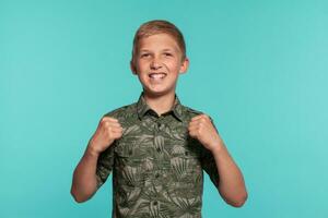 Close-up portrait of a blonde teenage boy in a green shirt with palm print posing against a blue studio background. Concept of sincere emotions. photo