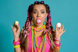 Lovely girl with a multi-colored braids hairstyle and bright make-up, posing in studio against a blue background, holding marshmallow in her hands. photo
