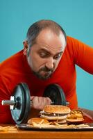 Close-up portrait of a middle-aged man with beard, dressed in a red turtleneck, posing with burgers and french fries. Blue background. Fast food. photo