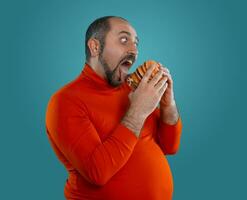 Close-up portrait of a middle-aged man with beard, dressed in a red turtleneck, posing with burgers against a blue background. Fast food. photo