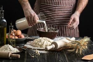 Baker in uniform pouring milk from plastic bottle into bowl. A handful of flour with egg on a rustic kitchen. photo