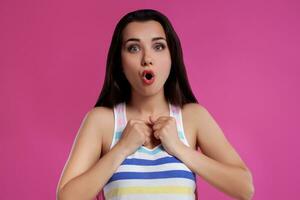 Brunette woman with long hair, dressed in colorful striped shirt, posing against pink studio background. Sincere emotions. Close-up. photo