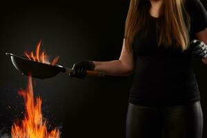 Brunette lady with tattooed hands, dressed in leggings and t-shirt. Holding a wok pan above fire against black background. Cooking concept. Side view photo