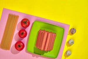 A lot of raw sausages on plate. On yellow background with pasta and vegetables, top view. photo