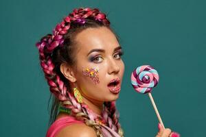 Lovely girl with a multi-colored braids hairstyle and bright make-up, posing in studio against a blue background, holding a lollipop in her hand. photo