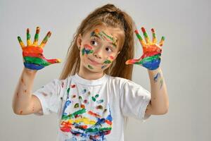 Little girl in white t-shirt is posing standing isolated on white and gesticulating with her painted in different colors palms. Art studio. Close-up. photo