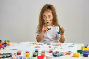 Little girl in white t-shirt sitting at table with whatman and paints on it, posing with painted face and hands. Isolated on white. Medium close-up. photo