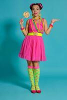 Lovely girl with a multi-colored braids hairstyle and bright make-up, posing in studio against a blue background, holding a lollipop in her hand. photo