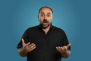 Close-up portrait of a brunet middle-aged man with beard, dressed in a black t-shirt and posing against a blue background. photo