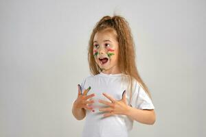 Little girl in white painted t-shirt is posing standing isolated on white and gesticulating with her colored in different paints palms and face. Art studio. Close-up. photo
