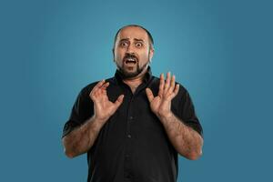 Close-up portrait of a brunet middle-aged man with beard, dressed in a black t-shirt and posing against a blue background. photo