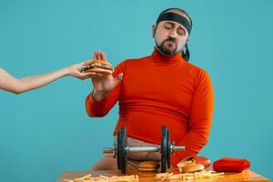 Middle-aged man with beard, dressed in a red turtleneck, headband, posing with burgers and french fries. Blue background. Close-up. Fast food. photo