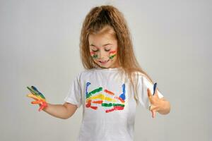 Little girl in white painted t-shirt is posing standing isolated on white and gesticulating with her colored in different paints palms and face. Art studio. Close-up. photo