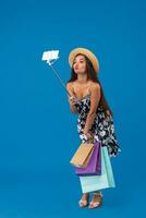 Young woman in casual clothes with shopping bags using selfie stick to take a self portrait on blue studio background with copy space photo