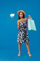 Young woman in casual clothes with shopping bags using selfie stick to take a self portrait on blue studio background with copy space photo