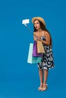 Young woman in casual clothes with shopping bags using selfie stick to take a self portrait on blue studio background with copy space photo