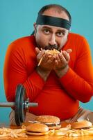 de edad mediana hombre con barba, vestido en un rojo suéter tipo con cuello de tortuga, venda, posando con hamburguesas y francés papas fritas azul antecedentes. de cerca. rápido alimento. foto