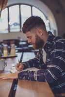 Young bearded businessman sits in cafe, home at table and writes photo