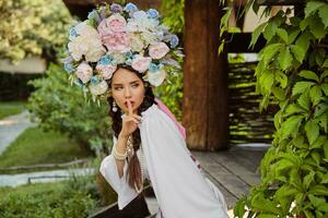 morena niña en un blanco ucranio auténtico nacional disfraz y un guirnalda de flores es posando en contra un verde patio trasero. de cerca. foto