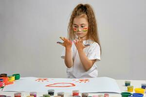 Little girl in white t-shirt sitting at table with whatman and colorful paints, painting on it with her hands. Isolated on white. Medium close-up. photo