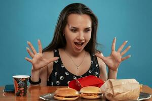 de cerca retrato de un mujer en un negro vestir posando sentado a el mesa con hamburguesas, francés papas fritas y beber. azul antecedentes. rápido alimento. foto